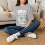 A woman sitting cross-legged on a woven rug in front of a beige sofa, wearing a heather gray graphic T-shirt with a white tiger face and the words 'TIGERS HAYSI'.