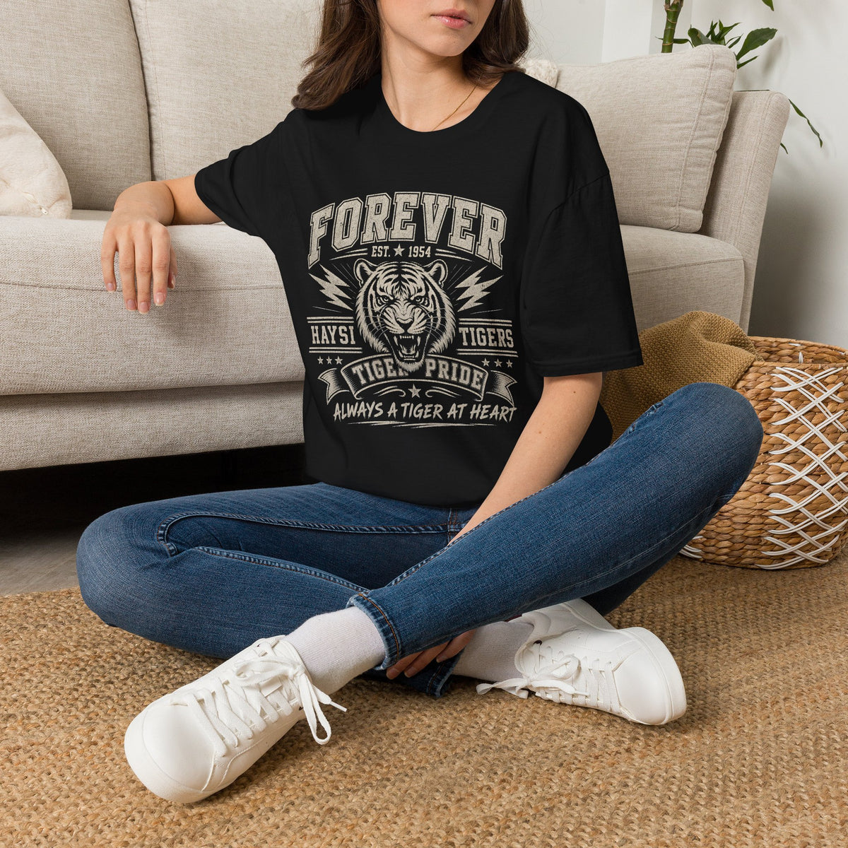 A woman wearing a black short-sleeve graphic t-shirt. The shirt features a white distressed print of a tiger head and the text 'FOREVER HAYSI TIGERS EST 1954 TIGRE PRIDE ALWAYS A TIGER AT HEART'.