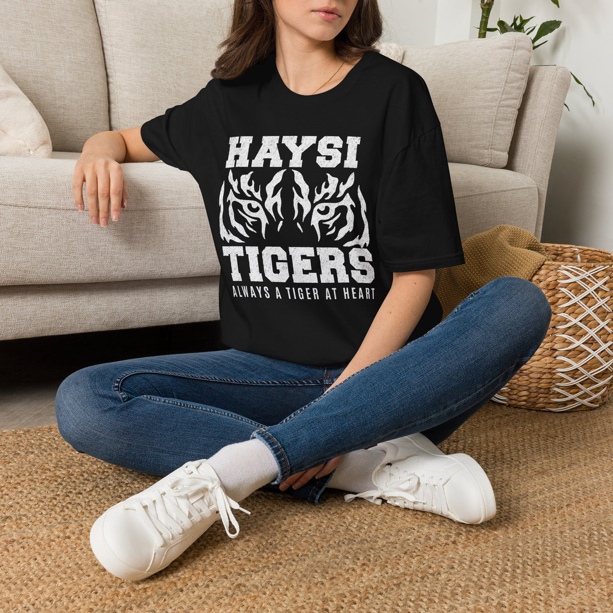 A woman wearing a black graphic T-shirt with white text reading 'HAYSI TIGERS' and a tiger illustration, blue jeans, and white sneakers.