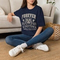 A woman sitting cross-legged on a woven rug in front of a beige sofa, wearing a navy blue graphic t-shirt with a distressed white and red 'Forever Tiger Pride' Tiger design, blue jeans, and white sneakers.