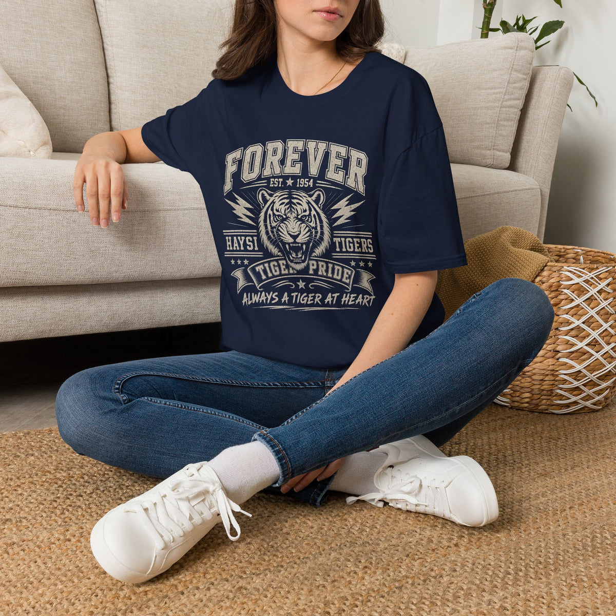 A woman sitting cross-legged on a woven rug in front of a beige sofa, wearing a navy blue graphic t-shirt with a distressed white and red 'Forever Tiger Pride' Tiger design, blue jeans, and white sneakers.