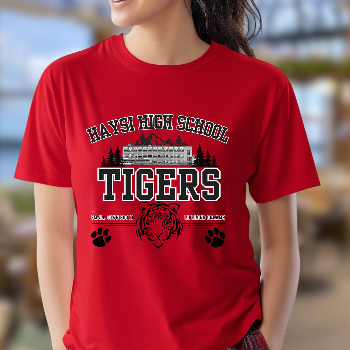 A woman wearing a bright red short-sleeve t-shirt with a black and white graphic that reads 'HAYSI HIGH SCHOOL TIGERS' with a tiger head, paw prints, and mountain/trees illustration, against a blurred outdoor background.