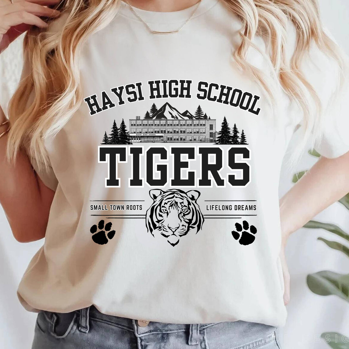 A woman wearing a cream-colored t-shirt with a black graphic print that reads 'HAYSI HIGH SCHOOL TIGERS' with a tiger head, mountains, paw prints, and 'SMALL TOWN ROOTS LIFELONG DREAMS'.