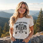 A woman standing outdoors in front of a mountain and forest landscape, wearing a cream-colored graphic t-shirt with 'HAYSI HIGH SCHOOL TIGERS' printed in black, along with a tiger head graphic and paw prints.