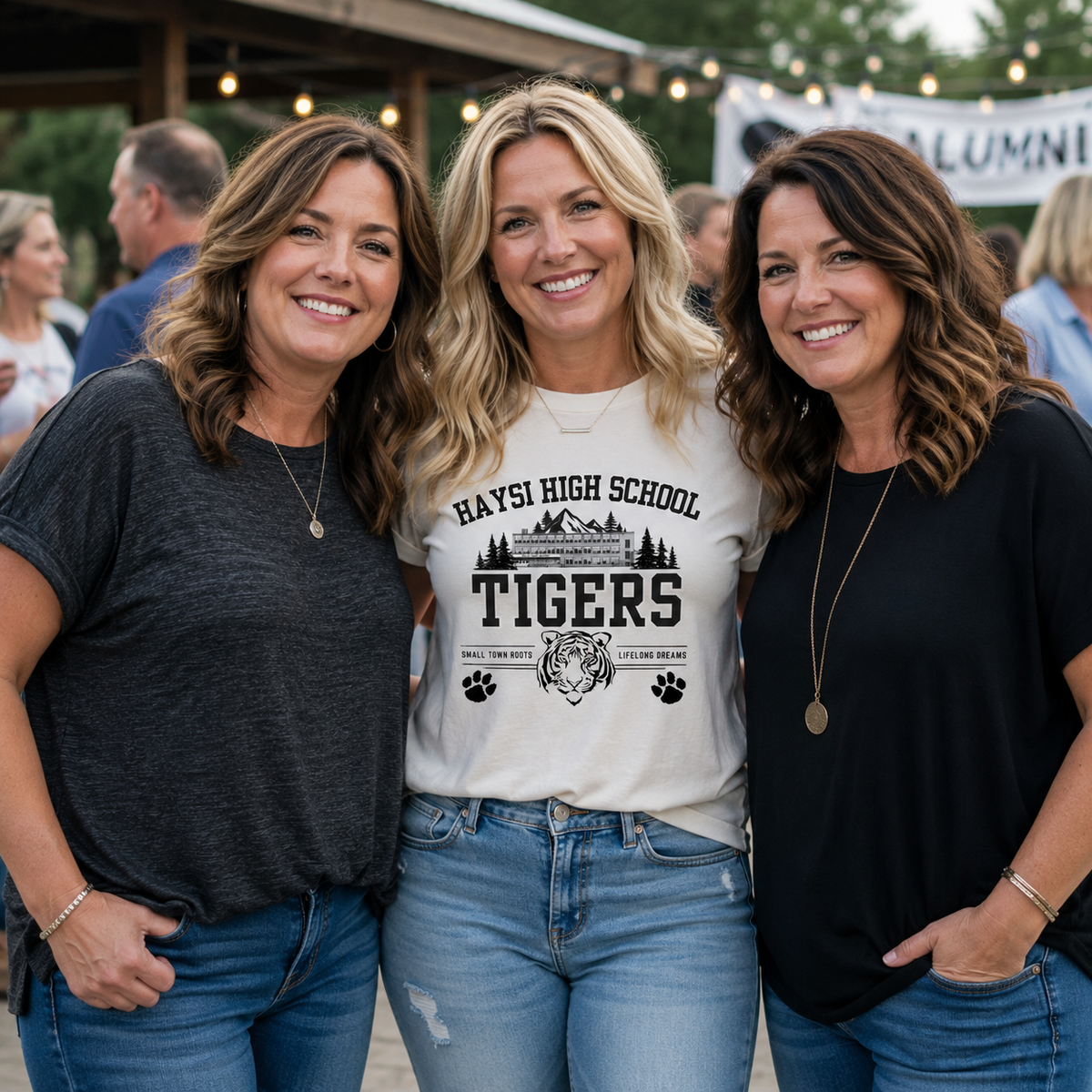 Three women smiling and posing together outdoors at what looks like a alumni or school event. The woman in the center is wearing a cream-colored graphic t-shirt with 'HAYS HIGH SCHOOL TIGERS' and a tiger graphic printed on the front, paired with blue jeans.