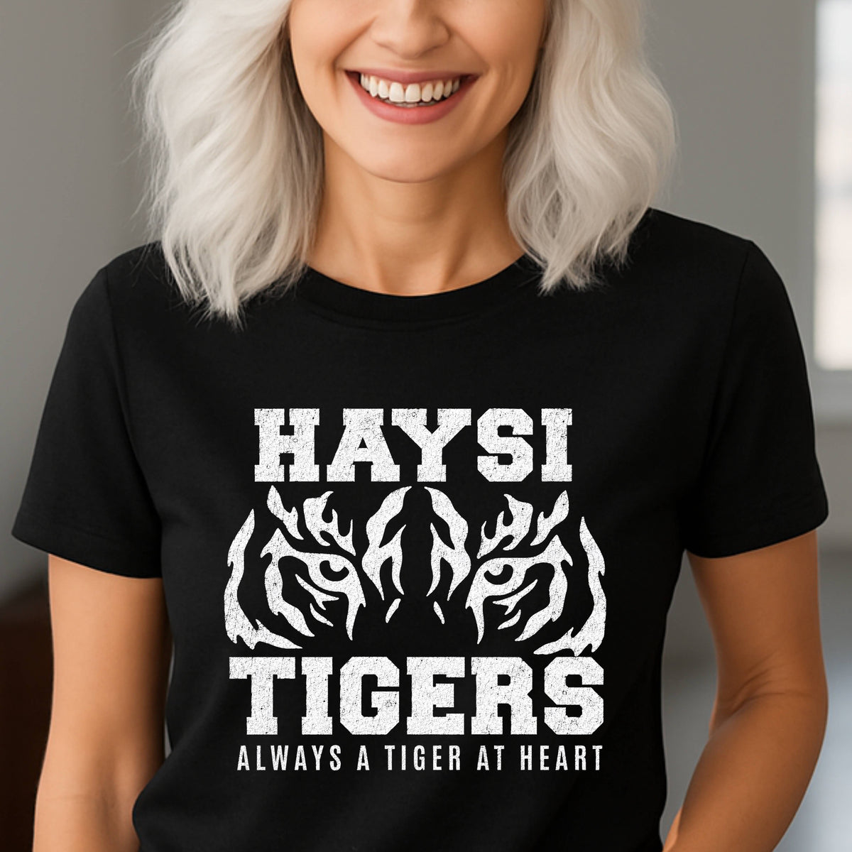 A black T-shirt with a large white graphic reading “HAYSI TIGERS” and “ALWAYS A TIGER AT HEART,” plus stylized tiger faces, against a softly blurred indoor background.