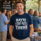 A man wearing a navy blue t-shirt with a white graphic that reads 'HAYSI TIGERS ALWAYS A TIGER AT HEART' stands at an outdoor reunion event with other people and string lights in the background.