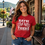 A red graphic T-shirt with white text reading 'HAYSI TIGERS' and 'ALWAYS A TIGER AT HEART,' paired with blue jeans. The background shows a blurred street scene with storefronts, a bench, plants, and hills.