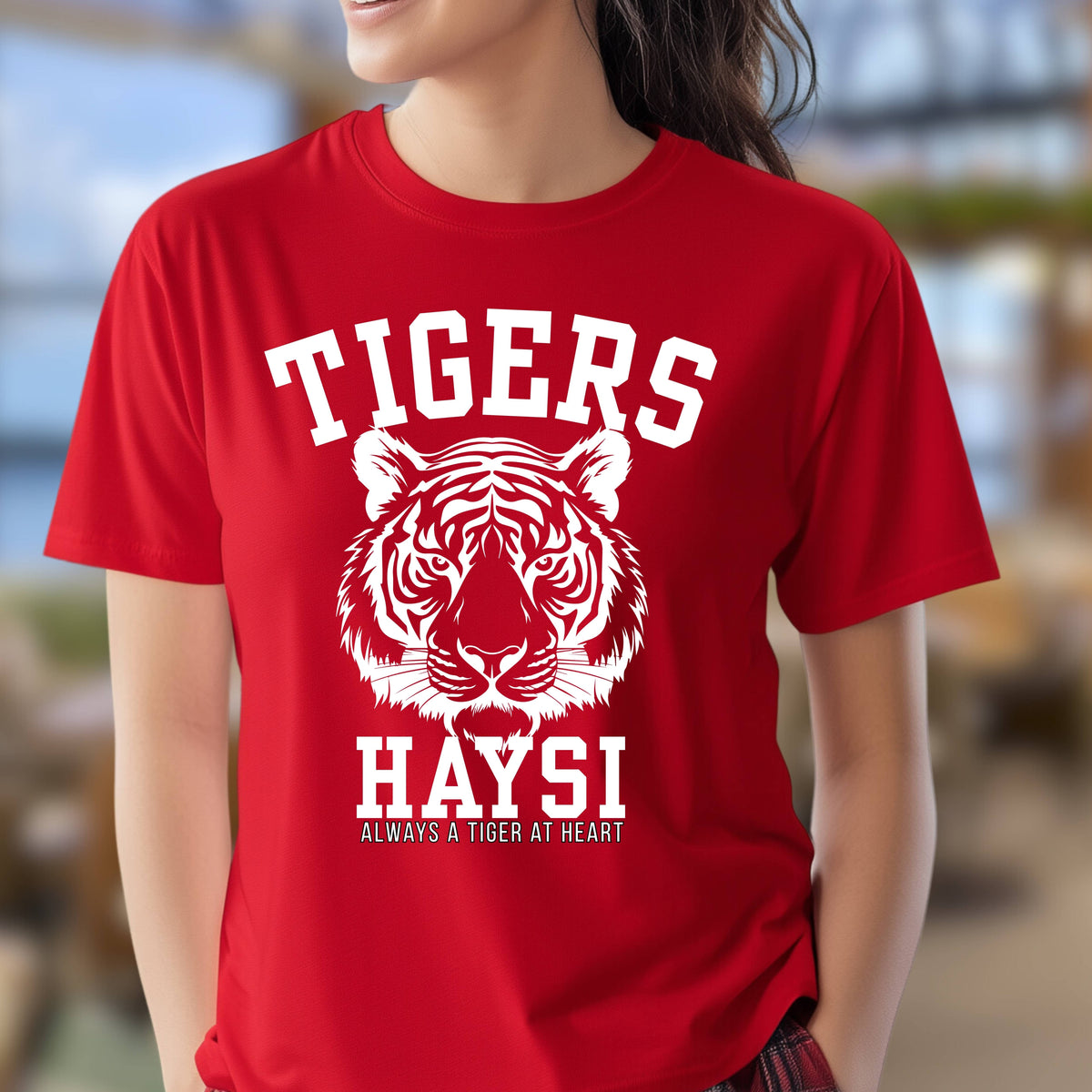 A woman wearing a bright red short-sleeve t-shirt with a white graphic print featuring a tiger head and the text 'TIGERS HAYSI ALWAYS A TIGER AT HEART'.