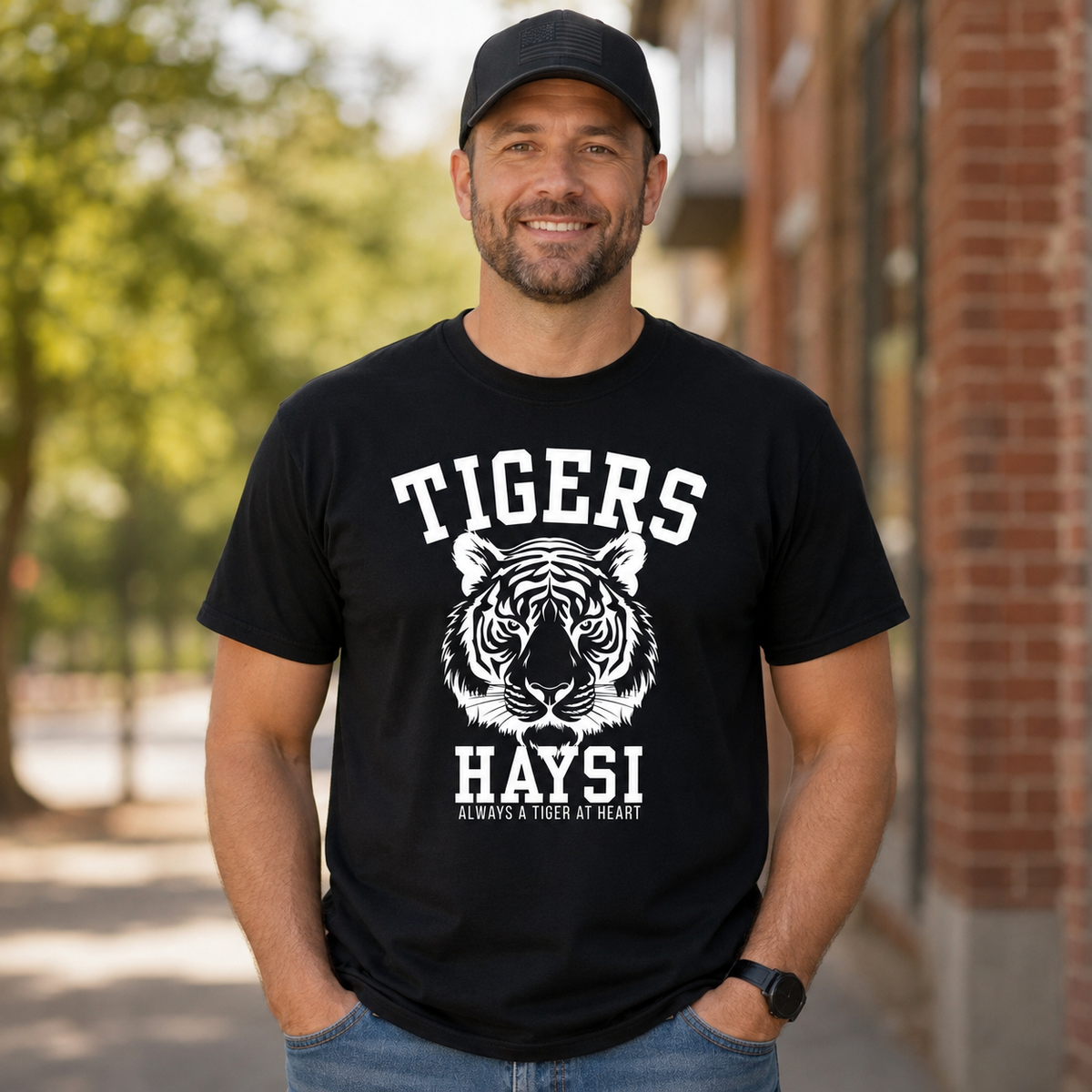 A man wearing a black crewneck t-shirt with a white graphic of a tiger head and the text 'TIGERS HAYSI ALWAYS A TIGER AT HEART', standing outdoors in front of a brick building.