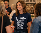 A woman smiling in a social setting, wearing a navy blue graphic t-shirt with a white tiger illustration and the text 'TIGERS HAYSI'.