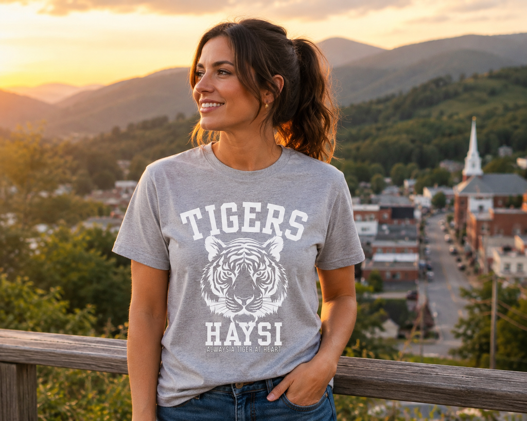A woman wearing a heather gray graphic t-shirt with a white tiger and 'TIGERS HAYSI' print, standing outdoors in front of a scenic mountain town at sunset.