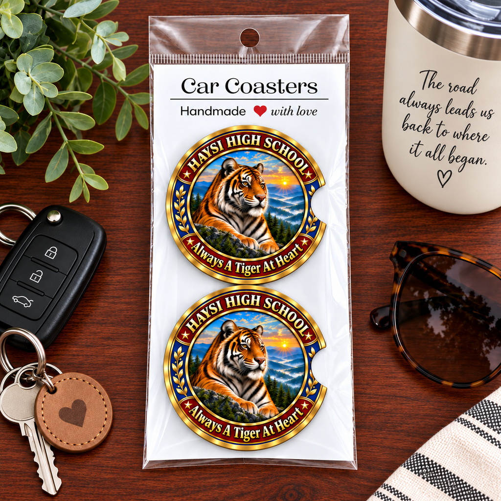 Two round car coasters featuring a tiger illustration and the text 'HAYS! HIGH SCHOOL' and 'Always A Tiger At Heart,' packaged in a clear plastic sleeve on a wooden tabletop with a key fob, mug, sunglasses, and greenery nearby.