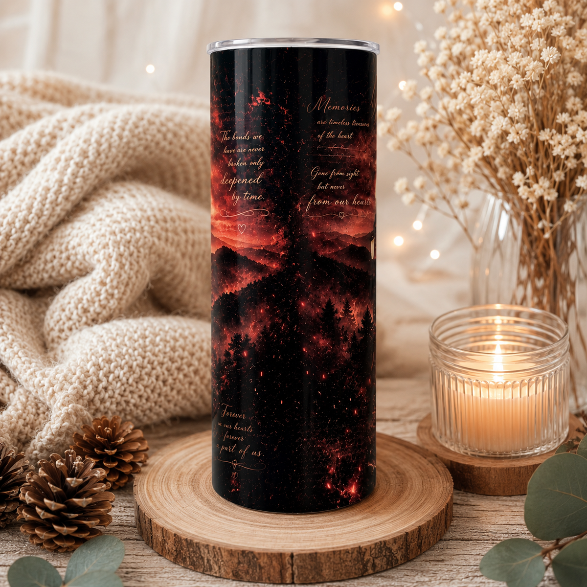 A tall insulated tumbler with a black background and a fiery red-orange landscape print, styled on a wooden slice with a knitted blanket, dried flowers, pinecones, and a lit candle in a cozy indoor setting.