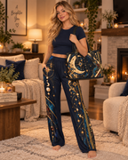 A woman stands barefoot in a cozy living room wearing a matching navy blue pajama set with gold celestial prints, including a short-sleeve top and wide-leg pants. She is holding a PU Leather shoulder bag with a moon and star pattern.